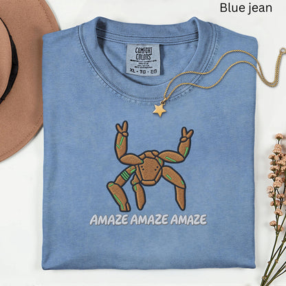May include: A blue jean colored t-shirt featuring an embroidered brown spider-like creature with green accents, making peace signs with two upper limbs. The text AMAZE AMAZE AMAZE is embroidered below the creature. The shirt has a Comfort Colors label.