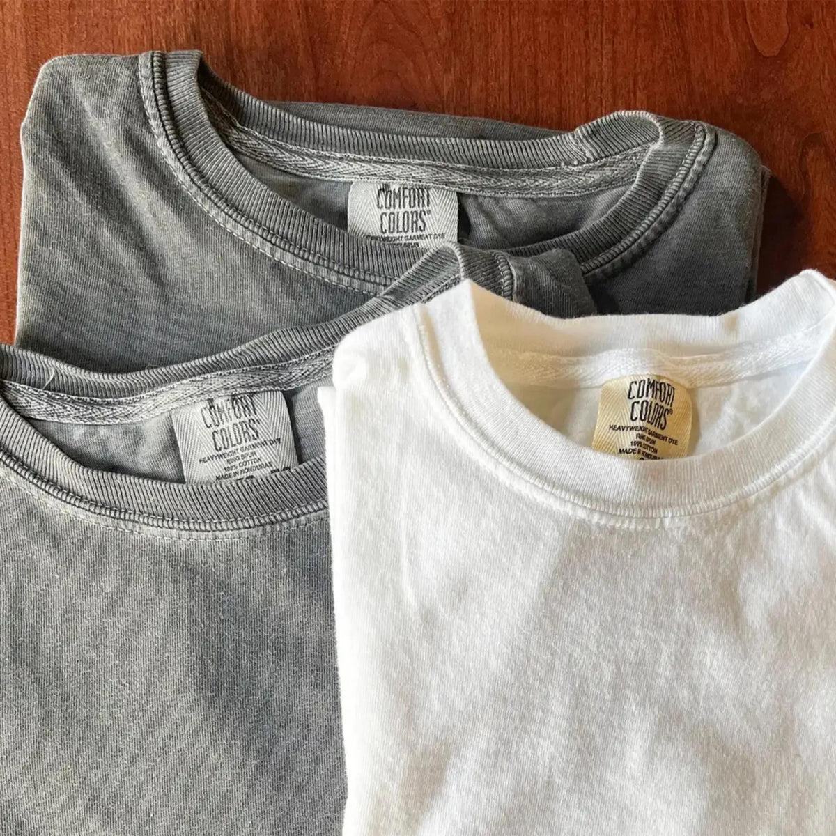 May include: Two crew neck t-shirts, one in a solid white and one in a washed-out charcoal gray. Each shirt features a ribbed collar and a white interior neck label with the text Comfort Colors, indicating the brand and garment details.