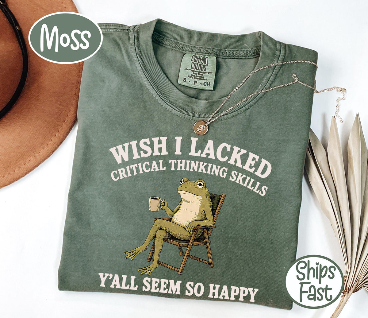May include: A moss green short-sleeve t-shirt featuring a graphic of a frog sitting in a folding chair holding a mug. The text reads WISH I LACKED CRITICAL THINKING SKILLS Y'ALL SEEM SO HAPPY. The shirt has a crew neckline and a Comfort Colors brand label.