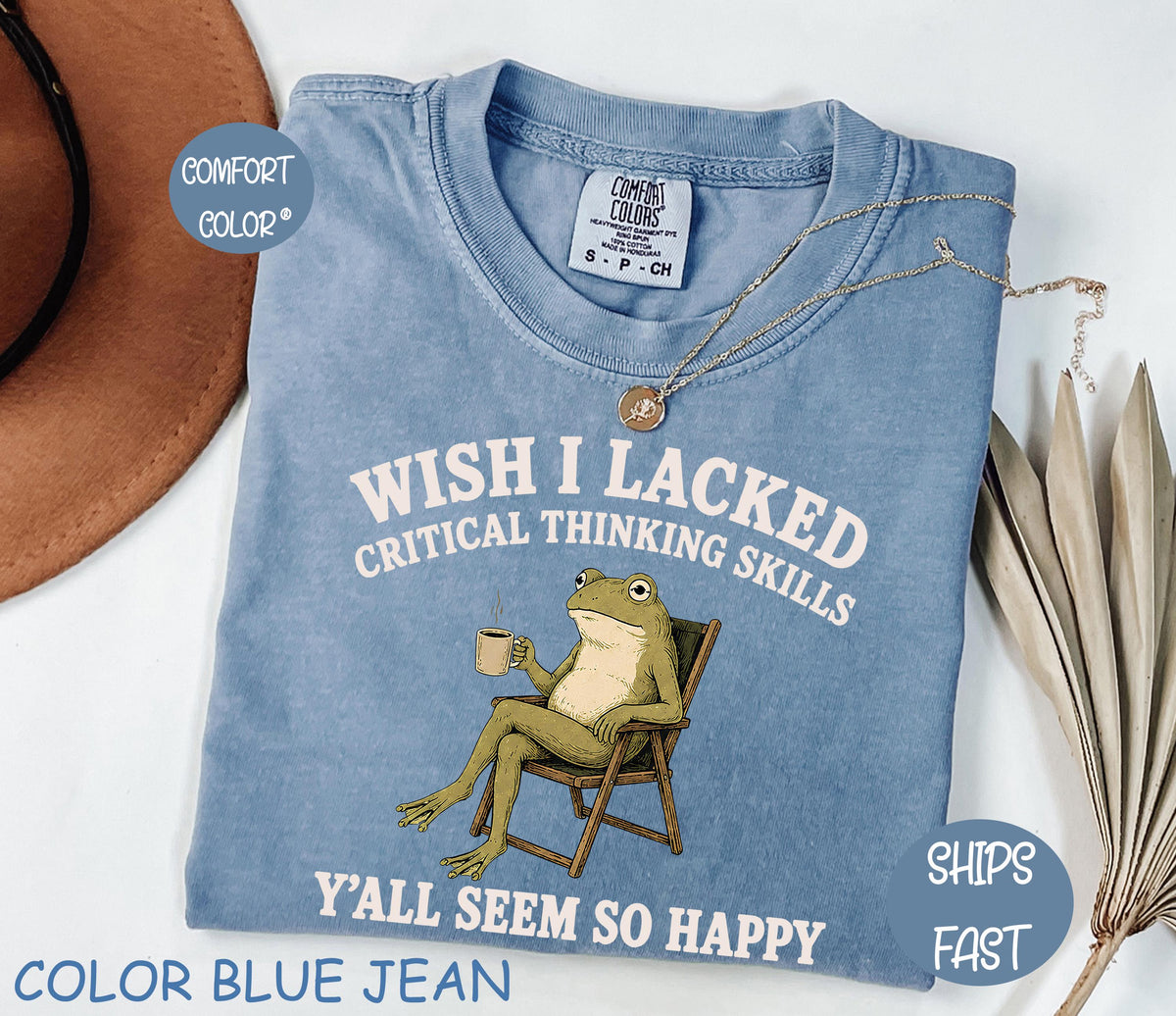May include: A blue jean colored t-shirt featuring a graphic of a green frog sitting in a wooden chair while holding a coffee mug. The text reads, "WISH I LACKED CRITICAL THINKING SKILLS Y'ALL SEEM SO HAPPY." The shirt has a Comfort Colors brand label.