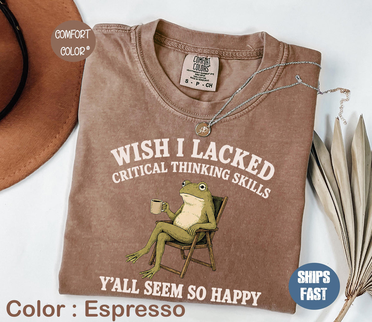 May include: An espresso-colored short-sleeve t-shirt featuring a graphic of a green frog sitting in a folding chair holding a mug. The text reads: WISH I LACKED CRITICAL THINKING SKILLS Y'ALL SEEM SO HAPPY. The shirt has a crew neckline and a tag.