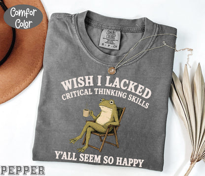 May include: A charcoal gray short-sleeve t-shirt featuring a graphic of a green frog sitting in a folding chair holding a coffee mug. The text reads "WISH I LACKED CRITICAL THINKING SKILLS Y'ALL SEEM SO HAPPY" in white lettering. The shirt has a crew neckline.