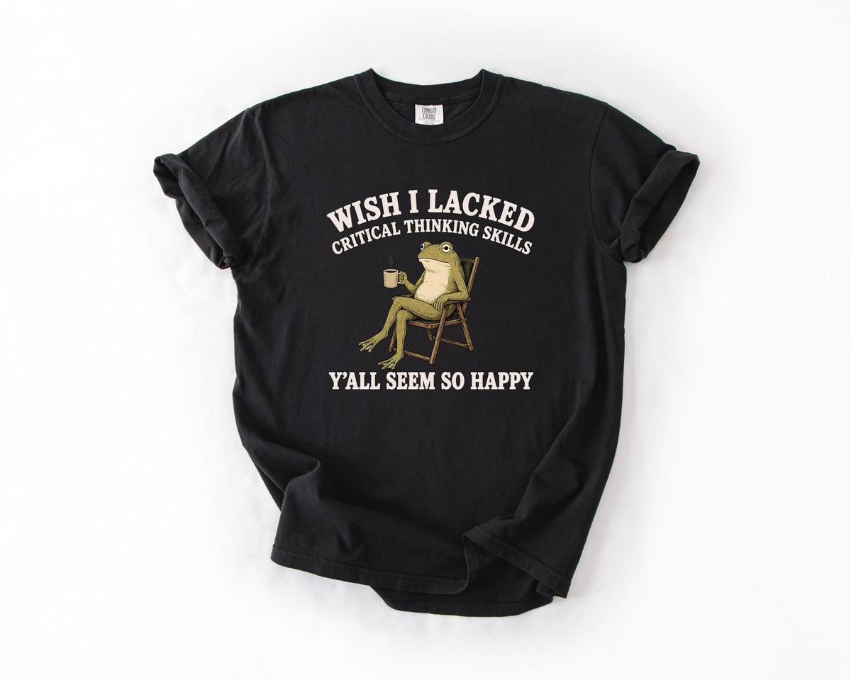 May include: A black short-sleeve t-shirt featuring a graphic of a green frog sitting in a folding chair holding a mug. The text above the frog reads WISH I LACKED CRITICAL THINKING SKILLS and the text below the frog reads Y'ALL SEEM SO HAPPY.