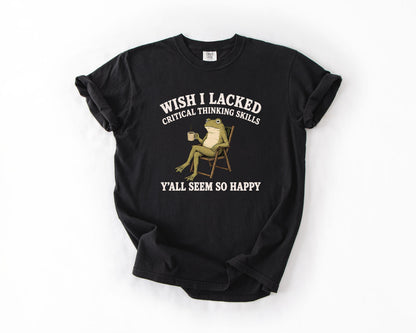 May include: A black short-sleeve t-shirt featuring a graphic of a green frog sitting in a folding chair holding a mug. The text above the frog reads WISH I LACKED CRITICAL THINKING SKILLS and the text below the frog reads Y'ALL SEEM SO HAPPY.