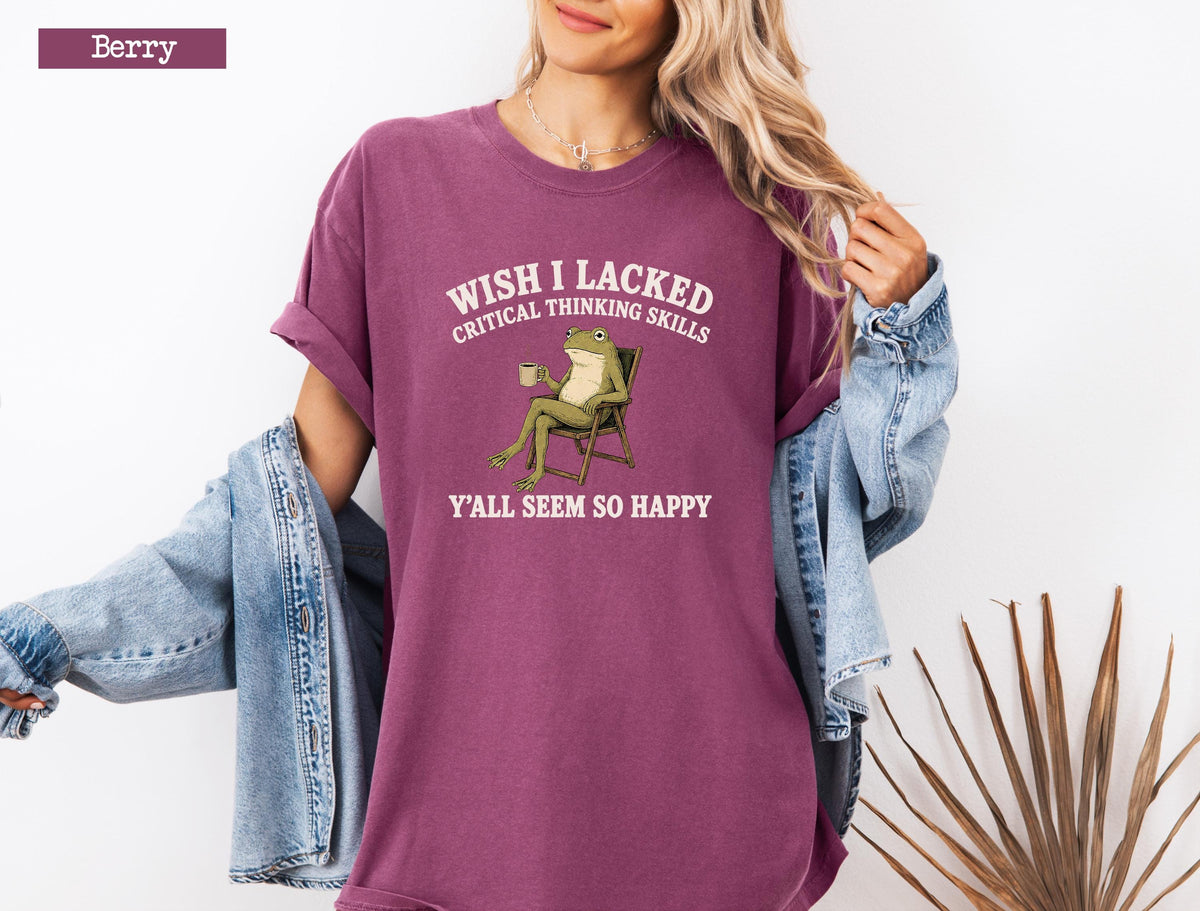 May include: A berry-colored short-sleeve t-shirt featuring a graphic of a green frog sitting in a chair holding a mug. The text above the frog reads WISH I LACKED CRITICAL THINKING SKILLS and the text below reads Y'ALL SEEM SO HAPPY in white.