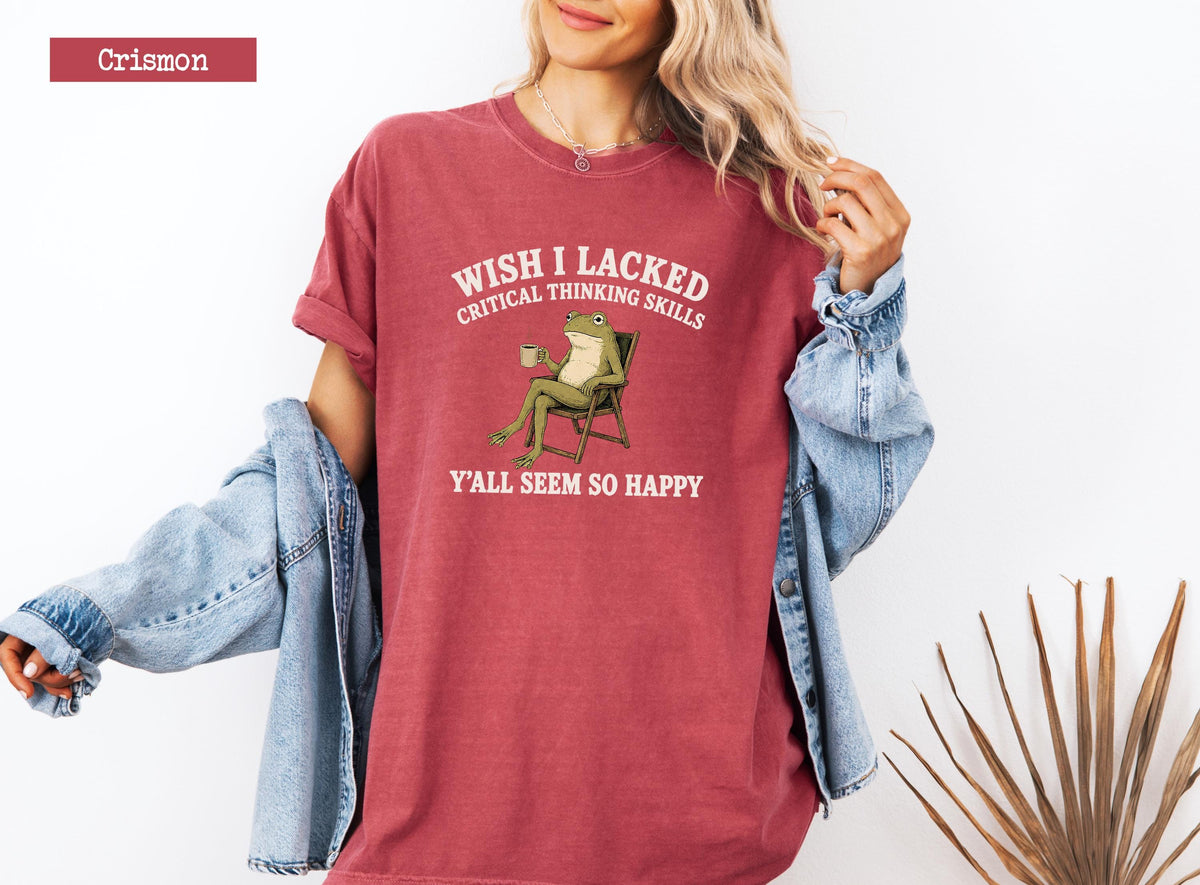 May include: A crimson crewneck t-shirt featuring a graphic of a green frog sitting in a folding chair while holding a mug. The text above the frog reads WISH I LACKED CRITICAL THINKING SKILLS and the text below reads Y'ALL SEEM SO HAPPY.
