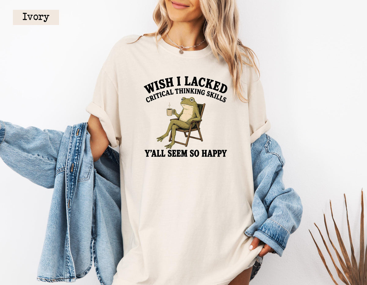 May include: An ivory short-sleeve t-shirt featuring a graphic of a green frog sitting in a chair holding a mug. The text above the frog reads WISH I LACKED CRITICAL THINKING SKILLS and the text below reads Y'ALL SEEM SO HAPPY. A blue denim jacket is draped over the arms.