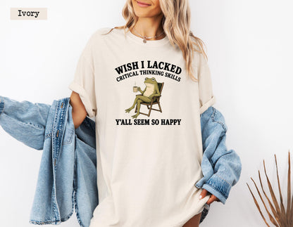 May include: An ivory short-sleeve t-shirt featuring a graphic of a green frog sitting in a chair holding a mug. The text above the frog reads WISH I LACKED CRITICAL THINKING SKILLS and the text below reads Y'ALL SEEM SO HAPPY. A blue denim jacket is draped over the arms.