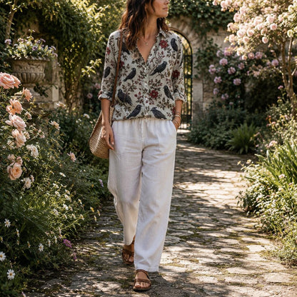 May include: A long-sleeved button-down shirt featuring a pattern of black crows and red flowers on a light gray background, paired with loose-fitting white trousers and a woven straw shoulder bag. The shirt has a collar and a front button placket.