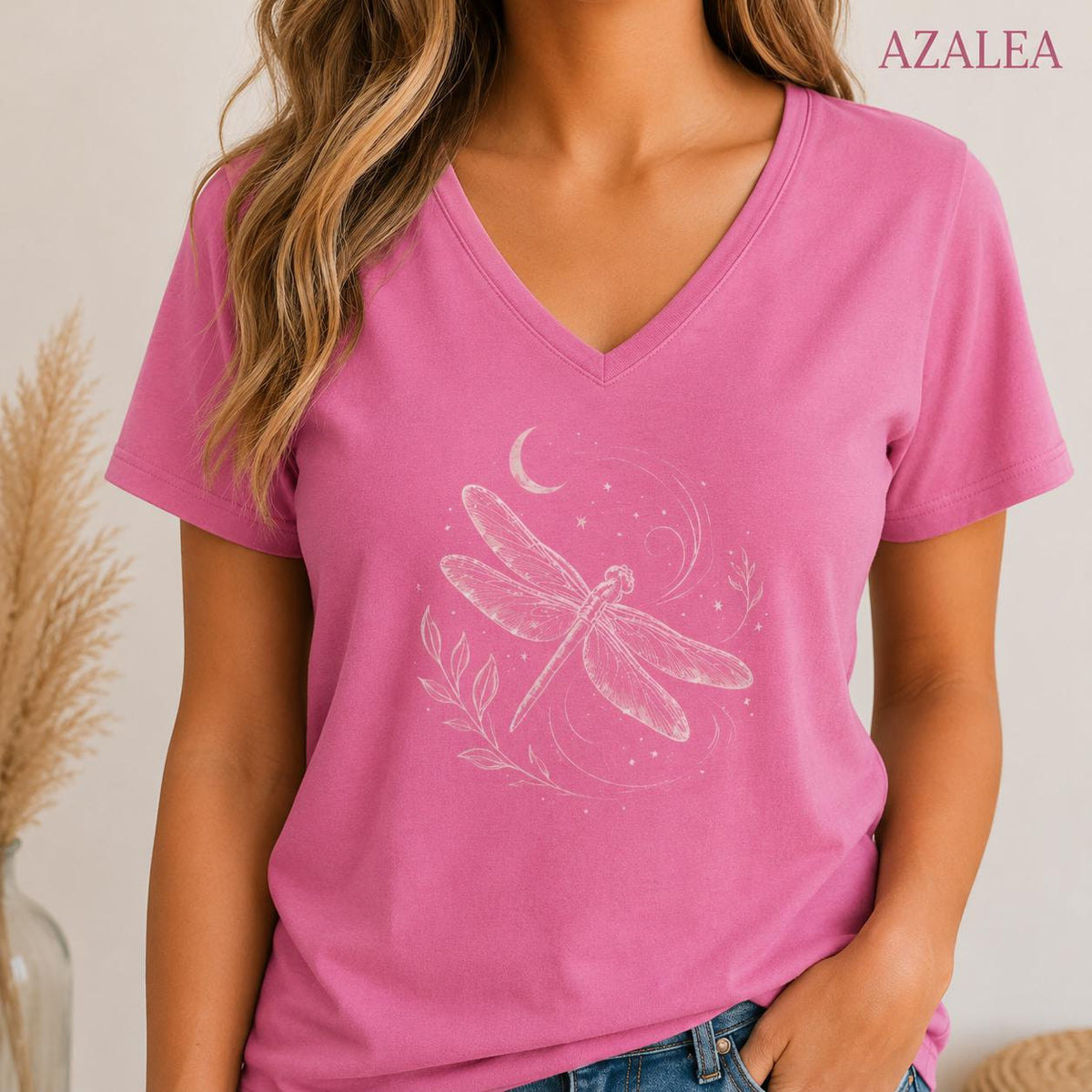 May include: A bright pink V-neck t-shirt featuring a white line art graphic of a dragonfly centered on the chest. The design includes a crescent moon, small stars, and delicate leafy sprigs surrounding the dragonfly. The word AZALEA is printed in the top right corner.