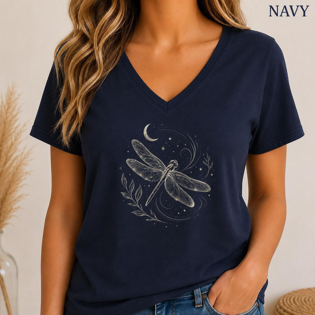 May include: A navy blue short-sleeve t-shirt with a V-neckline. The center of the shirt features a white graphic illustration of a dragonfly surrounded by swirling lines, small stars, a crescent moon, and leafy branches. The word NAVY is printed.