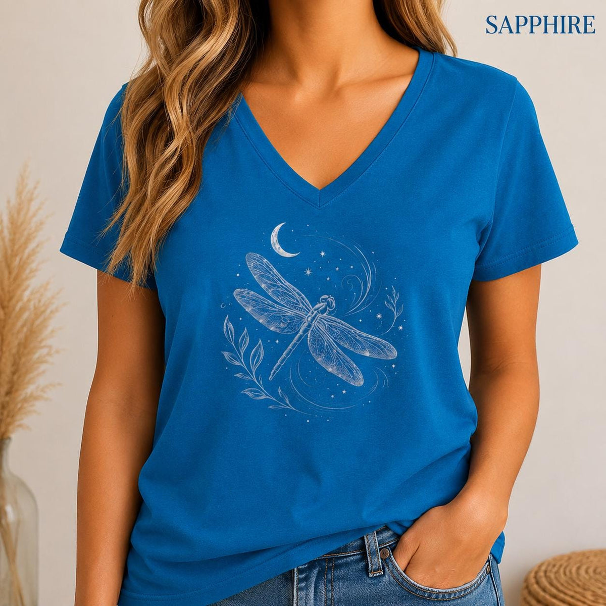 May include: A sapphire blue V-neck t-shirt featuring a white line art graphic of a dragonfly surrounded by a crescent moon, stars, and leafy sprigs. The word SAPPHIRE is printed in the upper right corner in a simple, clean, dark blue font.