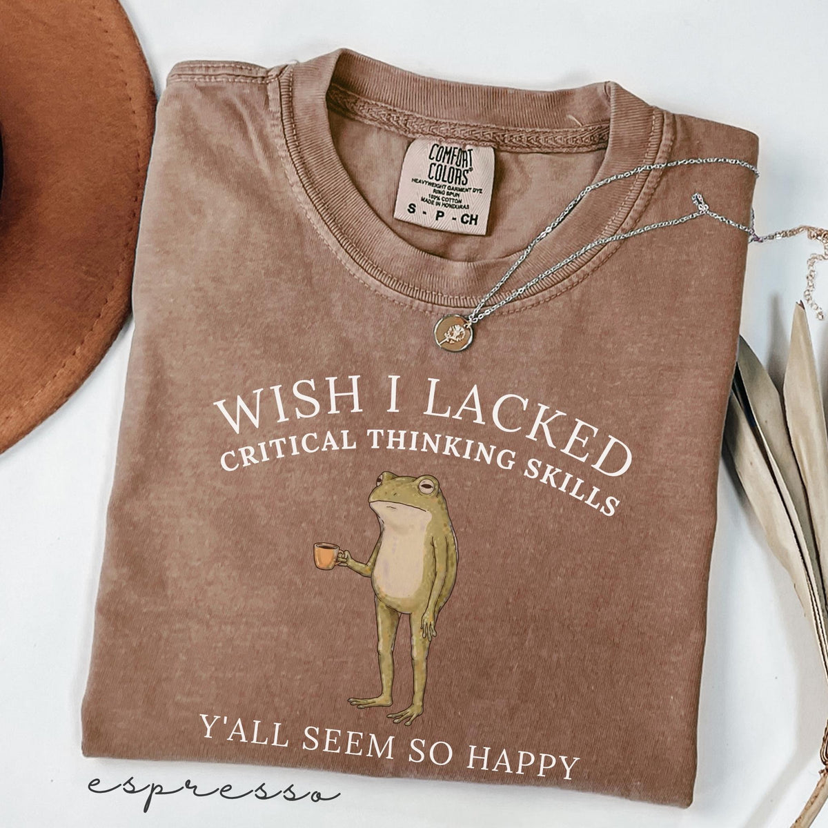 May include: A brown short-sleeved t-shirt featuring a graphic of a standing frog holding a small orange cup. The text reads WISH I LACKED CRITICAL THINKING SKILLS and Y'ALL SEEM SO HAPPY. The shirt has a Comfort Colors label in the neck area.