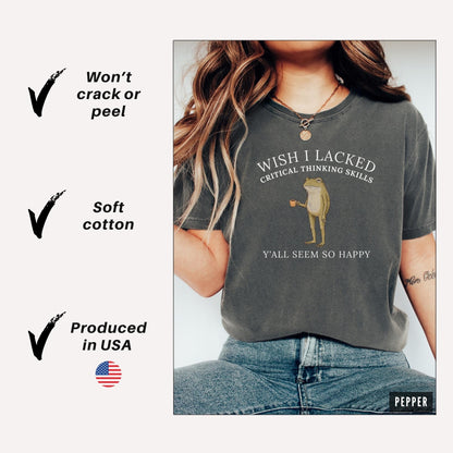 May include: A charcoal gray short-sleeve t-shirt featuring a graphic of a frog holding a small cup. The text reads WISH I LACKED CRITICAL THINKING SKILLS Y'ALL SEEM SO HAPPY. The shirt is displayed with text notes indicating it will not crack or peel.