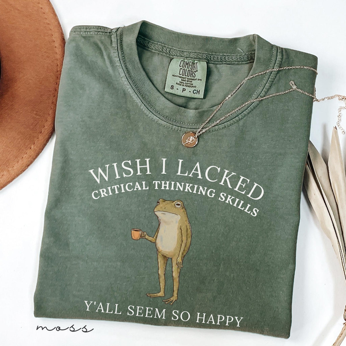 May include: A moss green crew neck t-shirt featuring a graphic of a standing frog holding a small orange mug. The text reads WISH I LACKED CRITICAL THINKING SKILLS Y'ALL SEEM SO HAPPY. The shirt has a Comfort Colors label at the inner neckline.