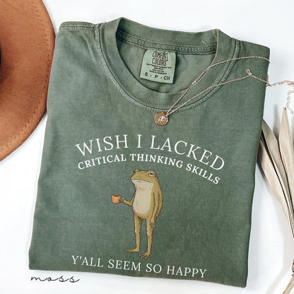 May include: A moss green crew neck t-shirt featuring a graphic of a standing frog holding a small orange mug. The text reads WISH I LACKED CRITICAL THINKING SKILLS Y'ALL SEEM SO HAPPY. The shirt has a Comfort Colors label at the inner neckline.