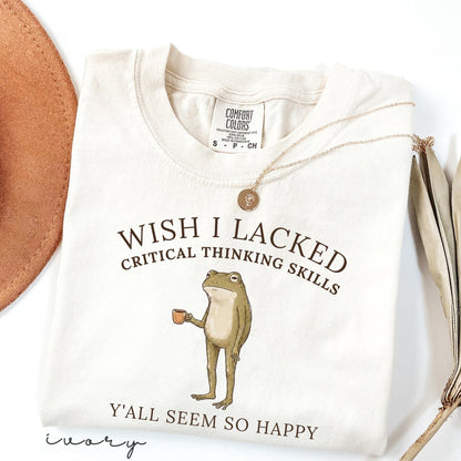 May include: An ivory crew neck t-shirt featuring a graphic of a standing frog holding a small brown mug. The text reads "WISH I LACKED CRITICAL THINKING SKILLS" above the frog and "Y'ALL SEEM SO HAPPY" below. The shirt has a Comfort Colors brand label.