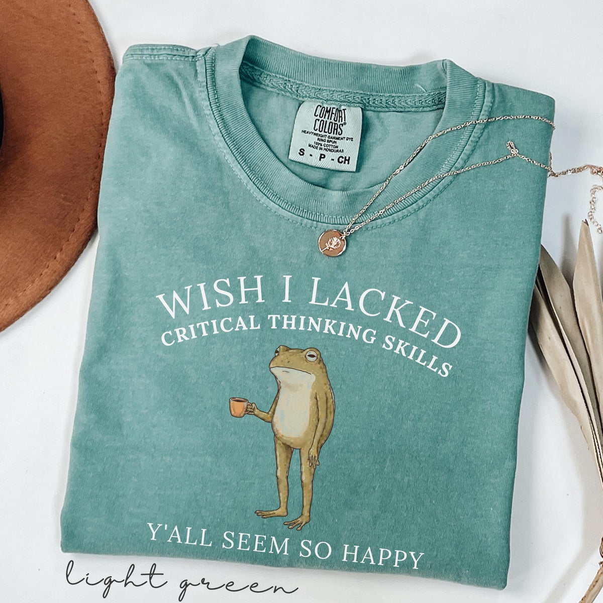 May include: A sage green short-sleeve t-shirt featuring a graphic of a standing frog holding a small orange mug. The text reads: WISH I LACKED CRITICAL THINKING SKILLS Y'ALL SEEM SO HAPPY. The shirt has a Comfort Colors brand label at the inner neckline.