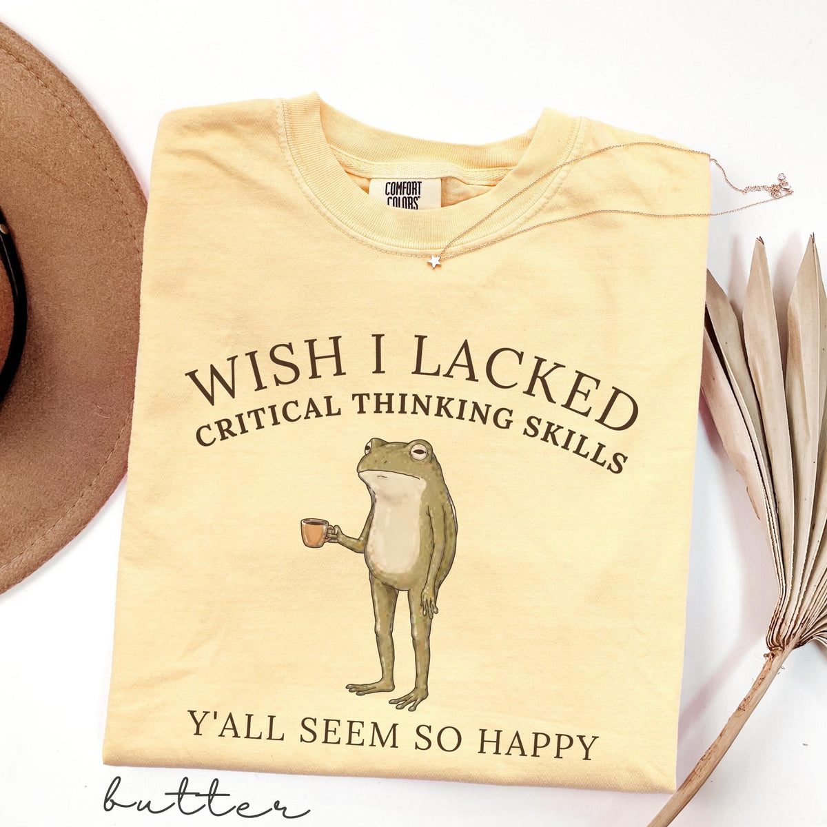 May include: A butter yellow short-sleeve t-shirt featuring a graphic of a standing frog holding a small brown mug. The text reads WISH I LACKED CRITICAL THINKING SKILLS Y'ALL SEEM SO HAPPY. The shirt has a Comfort Colors brand label at the inner neckline.
