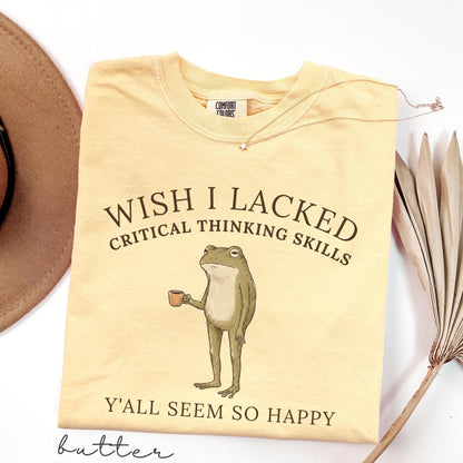 May include: A butter yellow short-sleeve t-shirt featuring a graphic of a standing frog holding a small brown mug. The text reads WISH I LACKED CRITICAL THINKING SKILLS Y'ALL SEEM SO HAPPY. The shirt has a Comfort Colors brand label at the inner neckline.