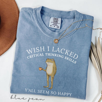 May include: A blue jean colored t-shirt featuring a graphic of a standing frog holding a small orange cup. The text reads WISH I LACKED CRITICAL THINKING SKILLS Y'ALL SEEM SO HAPPY. The shirt has a Comfort Colors brand label at the neck.