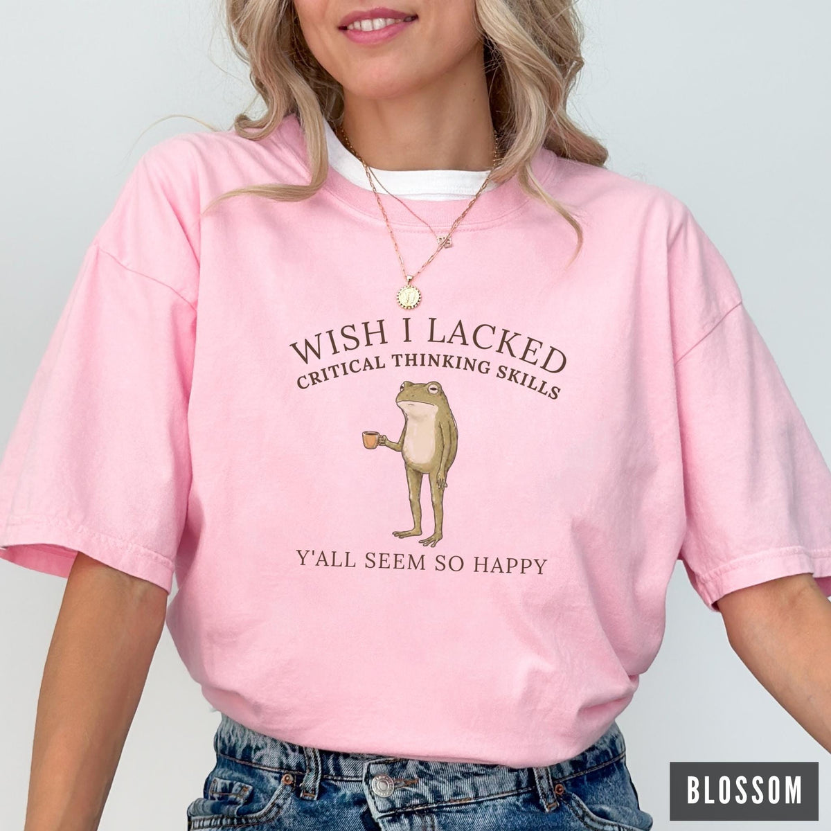 May include: A light pink short-sleeve t-shirt featuring a graphic of a standing frog holding a small brown cup. The text above the frog reads WISH I LACKED CRITICAL THINKING SKILLS and the text below reads Y'ALL SEEM SO HAPPY in dark brown font.