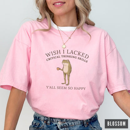 May include: A light pink short-sleeve t-shirt featuring a graphic of a standing frog holding a small brown cup. The text above the frog reads WISH I LACKED CRITICAL THINKING SKILLS and the text below reads Y'ALL SEEM SO HAPPY in dark brown font.
