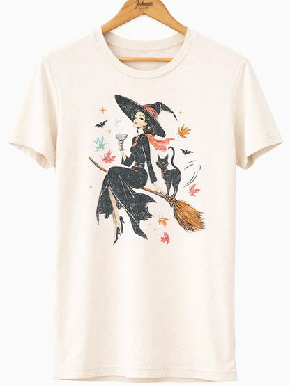 May include: A cream-colored short-sleeve t-shirt featuring a graphic print of a witch in a black dress and pointed hat sitting on a broomstick, holding a glass, accompanied by a black cat, small bats, and scattered autumn leaves in orange and red.