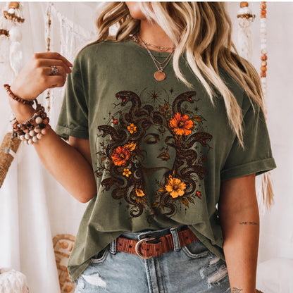 May include: An olive green short-sleeve t-shirt featuring a graphic print of a face surrounded by snakes and orange flowers. The design includes intricate line work and celestial elements. The shirt is paired with a brown leather belt and blue denim.