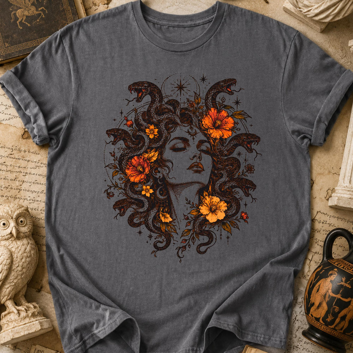 May include: A charcoal gray t-shirt featuring a detailed graphic print of a face surrounded by intertwined snakes and vibrant orange flowers. The illustration includes celestial star motifs and fine line work, centered on the chest of the garment.