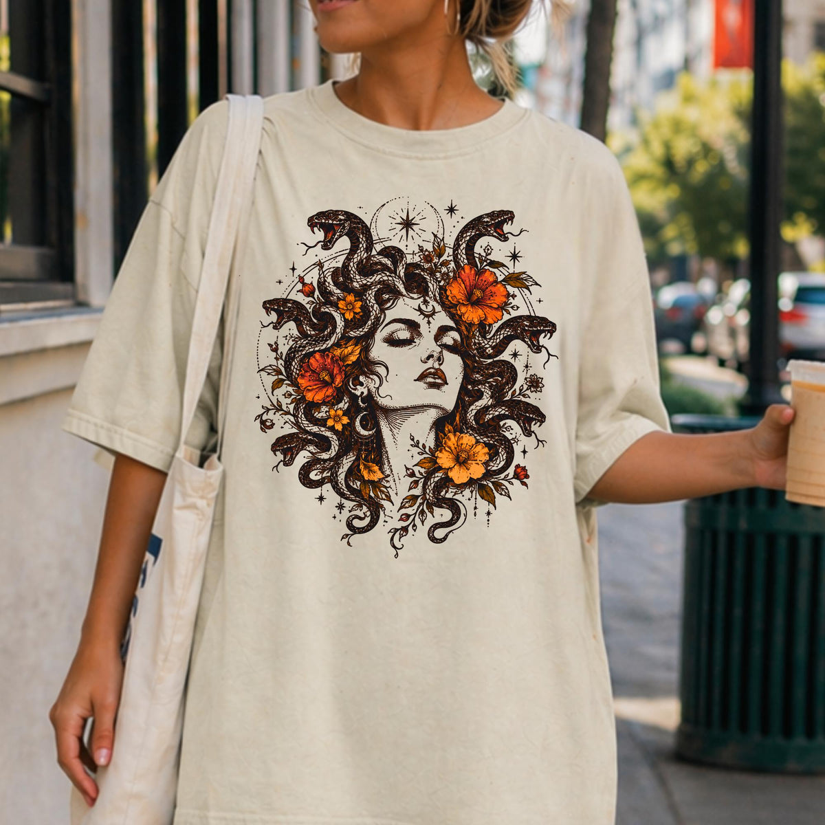 May include: An off-white short-sleeve t-shirt featuring a detailed graphic print of a person with snakes for hair, accented by orange flowers and celestial star symbols. The design is centered on the chest, showcasing a dark ink-style illustration.