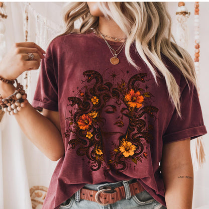 May include: A maroon short-sleeve t-shirt featuring a graphic print of a face surrounded by snakes and orange flowers. The design includes celestial star motifs and fine line work. The shirt is worn with a brown leather belt and layered necklaces.
