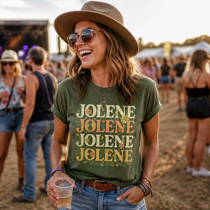 May include: An olive green short-sleeve t-shirt featuring the word JOLENE printed four times in a stacked, retro-style font with small floral illustrations integrated into the lettering, worn with a brown leather belt and layered bracelets.