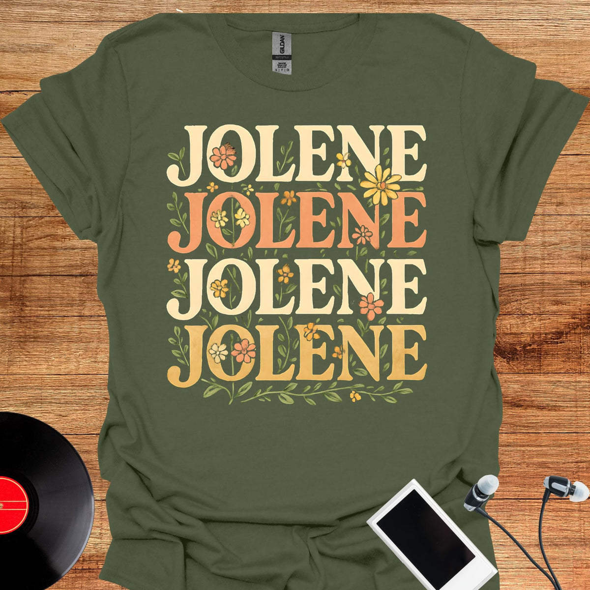 May include: An olive green short-sleeved t-shirt featuring the word JOLENE printed four times in a stacked layout. The text is rendered in a bold, rounded font with a color gradient from cream to orange and yellow, accented by small floral illustrations.