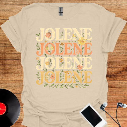 May include: A beige short-sleeve t-shirt featuring the name JOLENE printed four times in a stacked layout. The text alternates between cream and orange colors, decorated with small yellow and orange floral illustrations and green leaves.