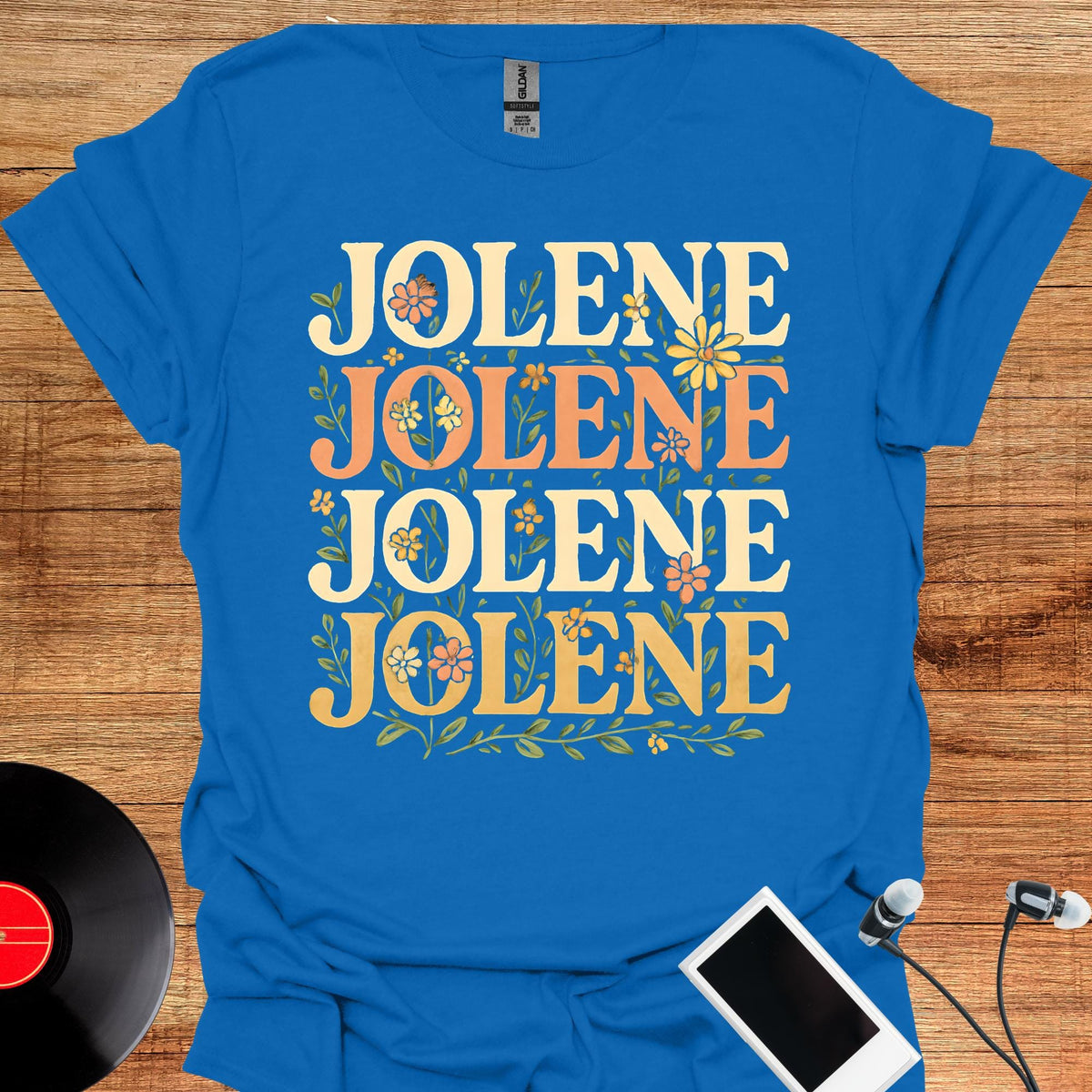 May include: A bright blue short-sleeved t-shirt featuring the name JOLENE printed four times in a stacked layout. The text is rendered in a cream-colored serif font and is decorated with small, colorful floral illustrations and green leaves.