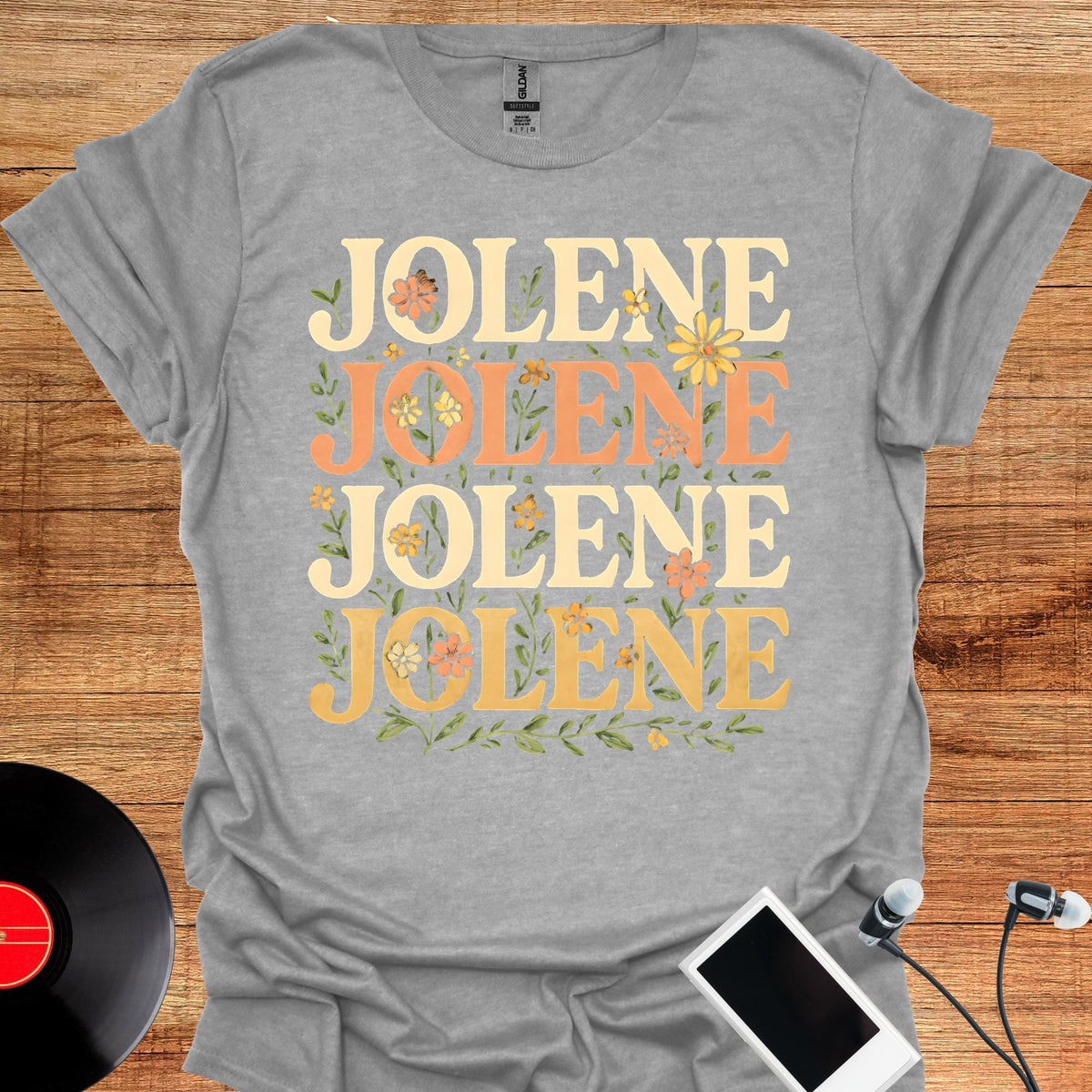 May include: A heather gray short-sleeve t-shirt featuring the name JOLENE printed four times in a stacked layout. The text alternates between cream and orange colors, decorated with small yellow and orange flowers and green leaves.