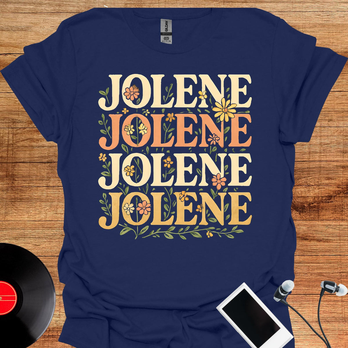 May include: A navy blue short-sleeve t-shirt featuring the word JOLENE printed four times in a stacked layout. The text is rendered in a cream and light orange font, decorated with small illustrations of yellow and orange flowers and green leaves.
