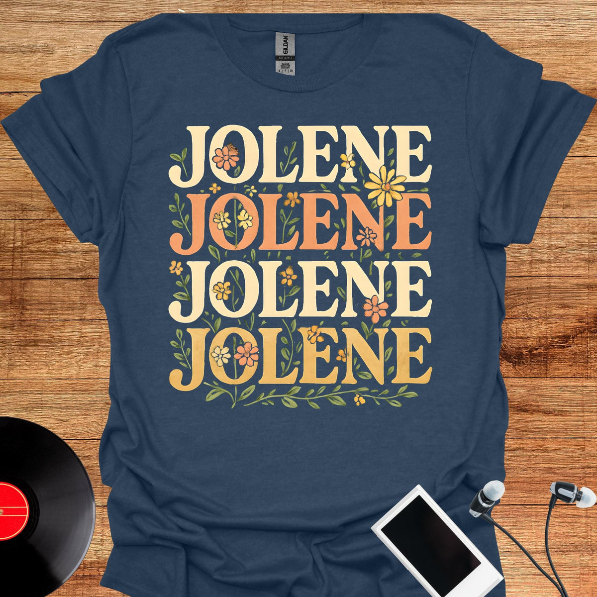 May include: A navy blue short-sleeved t-shirt featuring the word JOLENE printed four times in a stacked arrangement. The text is rendered in a bold, retro-style font in shades of cream and orange, accented with small green floral illustrations.