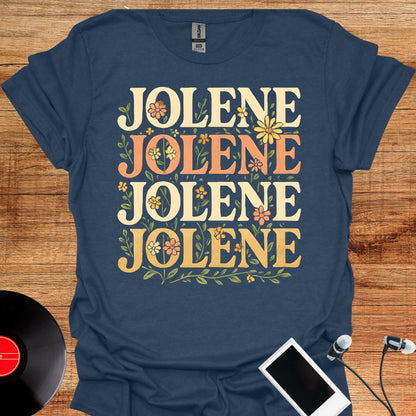 May include: A navy blue short-sleeved t-shirt featuring the word JOLENE printed four times in a stacked arrangement. The text is rendered in a bold, retro-style font in shades of cream and orange, accented with small green floral illustrations.