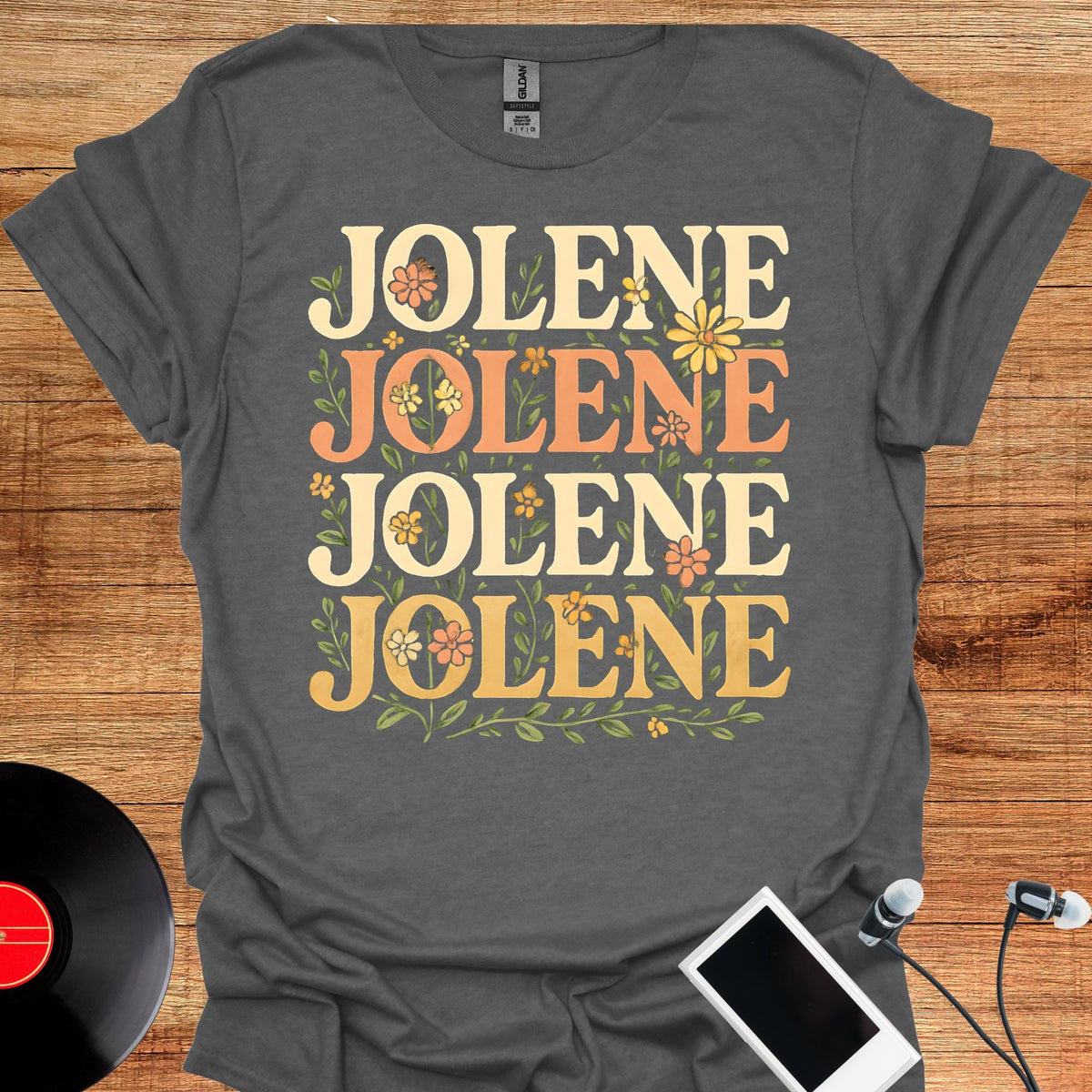 May include: A dark gray short-sleeve t-shirt featuring the name JOLENE printed four times in a stacked layout. The text is rendered in a bold, serif font with alternating cream and orange colors, surrounded by small green leaves and flowers.