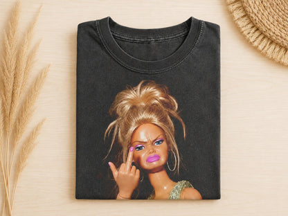 May include: A black crew neck t-shirt featuring a graphic print of a blonde doll with a messy bun hairstyle, wearing hoop earrings and a glittery top. The doll has a frowning expression and is holding up a middle finger with a pink fingernail.