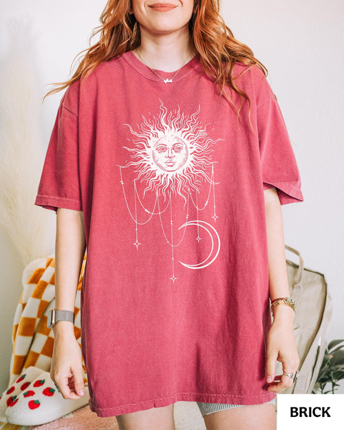 May include: A brick red short-sleeved t-shirt featuring a large white graphic print on the front. The design depicts a stylized sun with a human face, surrounded by radiating lines, hanging beaded chains, small stars, and a crescent moon.