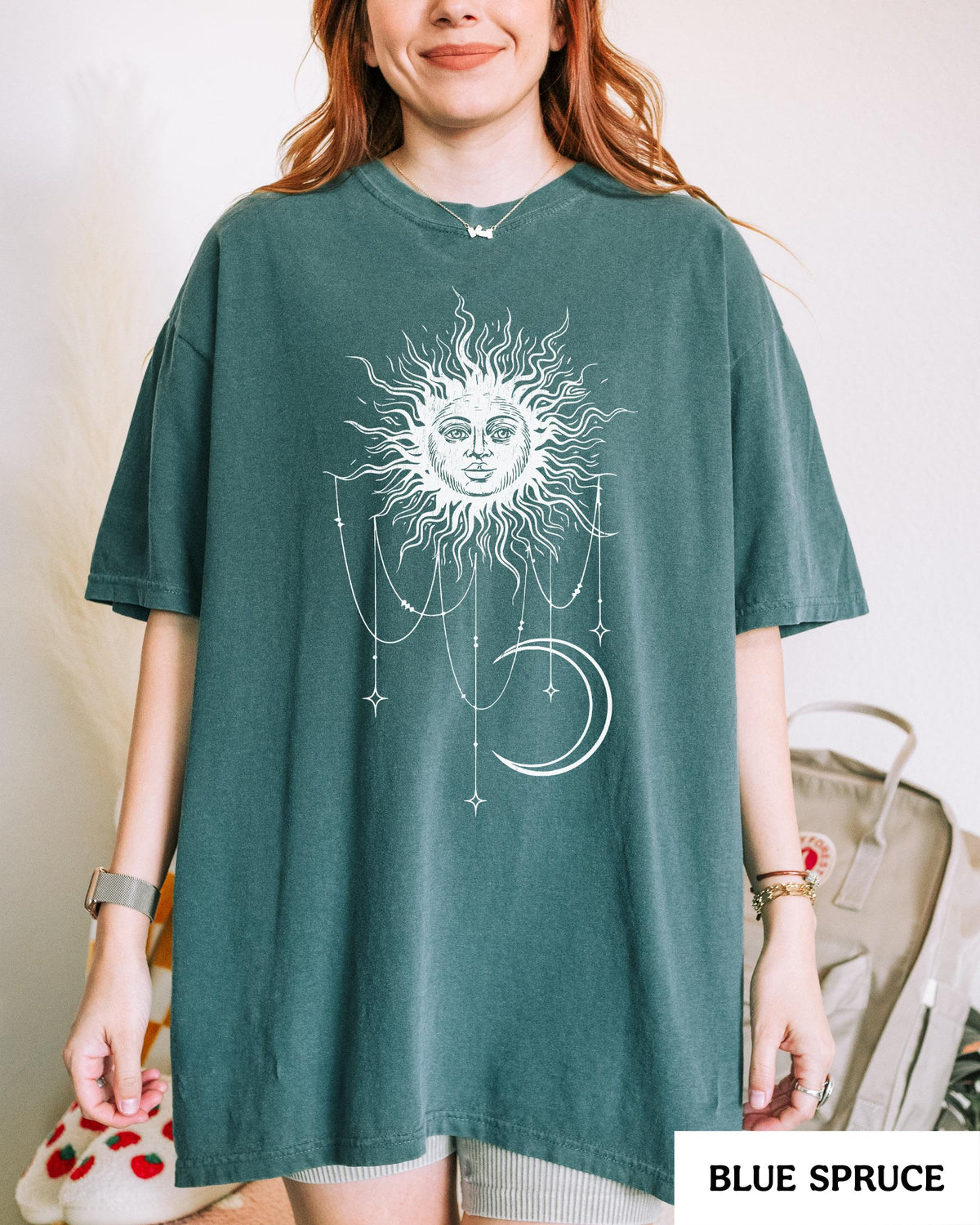 May include: A blue spruce colored short-sleeve t-shirt featuring a white graphic print on the front. The design depicts a sun with a human face surrounded by radiating rays, with hanging chains, stars, and a crescent moon positioned below it.