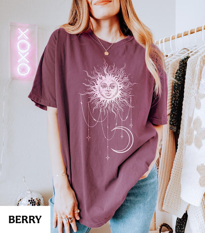 May include: A berry-colored short-sleeve t-shirt featuring a white graphic print of a sun with a face, a crescent moon, and hanging star-adorned chains. The shirt has a crew neckline and a relaxed fit, showcasing a celestial design on the front.