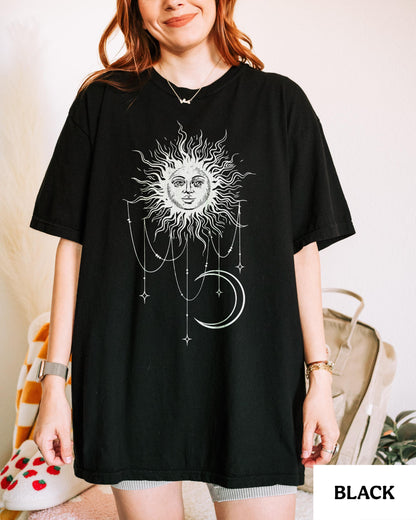 May include: A black short-sleeved t-shirt featuring a white graphic print of a sun with a human face, surrounded by dangling celestial chains and a crescent moon. The design is centered on the chest of the garment, which has a crew neckline.