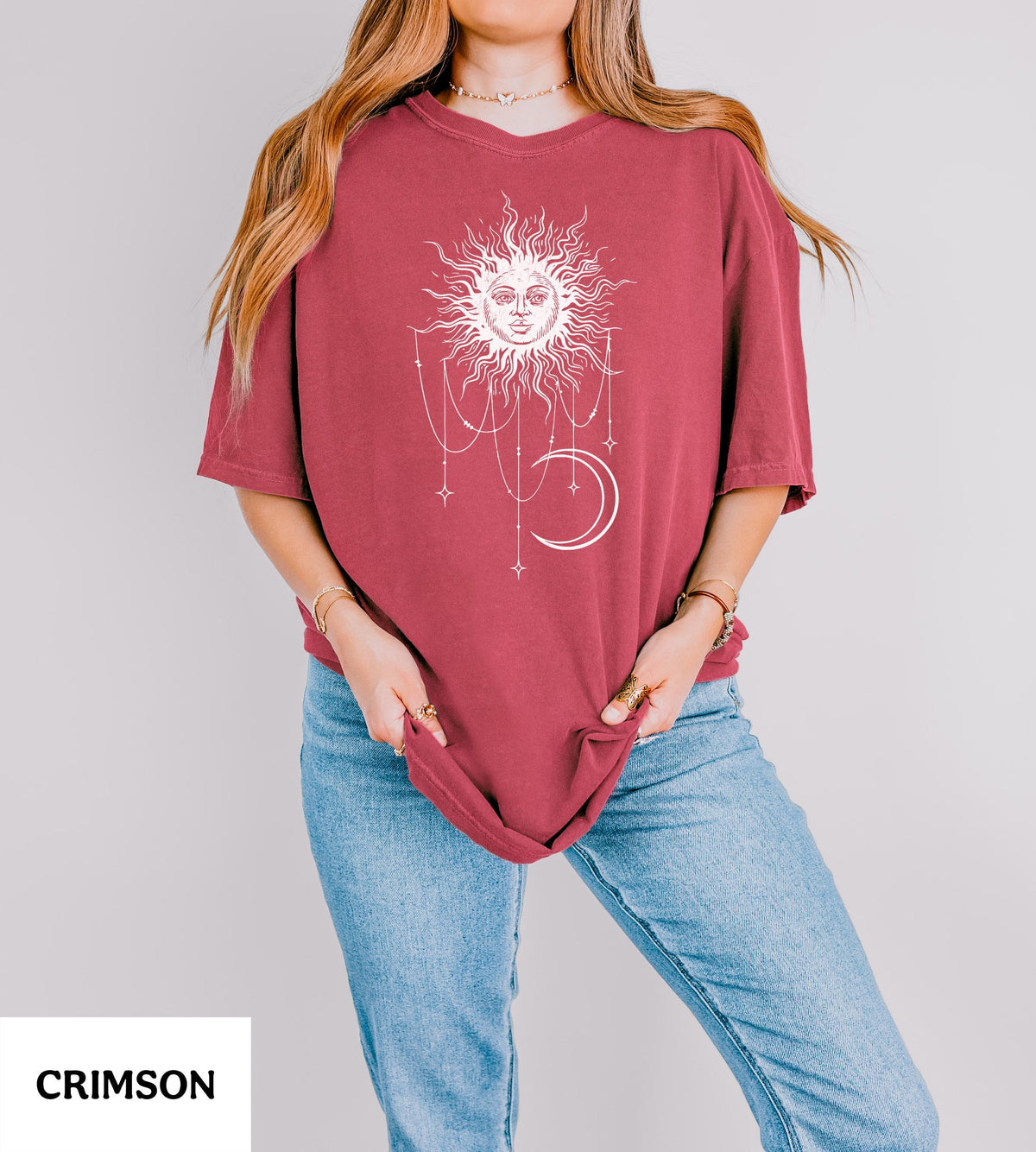 May include: A crimson short-sleeved t-shirt featuring a white graphic print of a sun with a human face, surrounded by hanging stars and a crescent moon. The shirt has a crew neckline and a loose, relaxed fit, paired with light blue denim jeans.