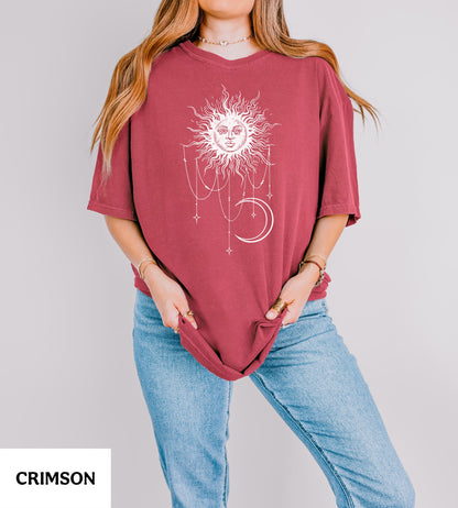 May include: A crimson short-sleeved t-shirt featuring a white graphic print of a sun with a human face, surrounded by hanging stars and a crescent moon. The shirt has a crew neckline and a loose, relaxed fit, paired with light blue denim jeans.
