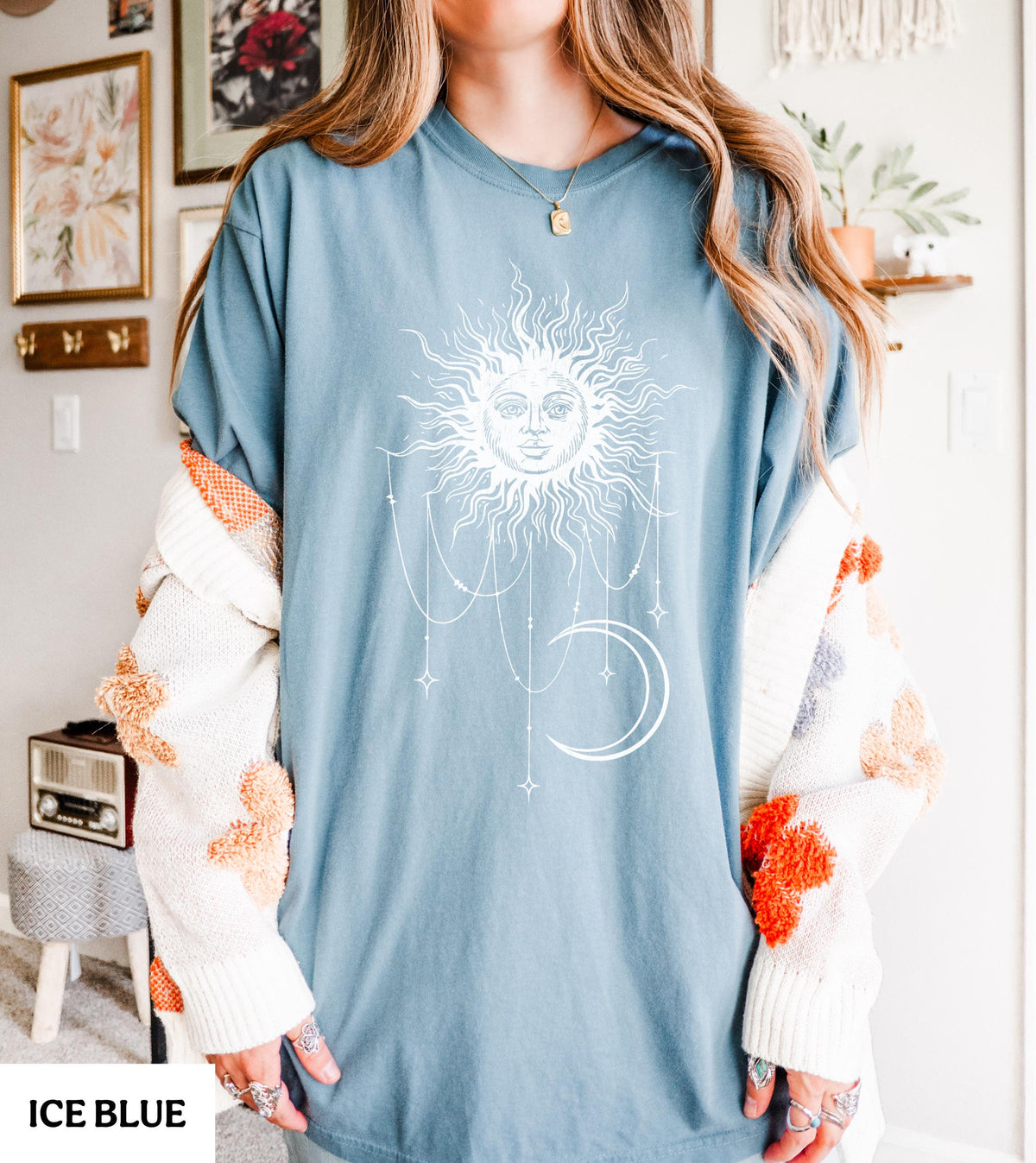 May include: An ice blue t-shirt featuring a white graphic print of a sun with a human face, surrounded by radiating rays, hanging celestial chains, and a crescent moon. The shirt has a crew neckline and short sleeves, worn with a white cardigan.
