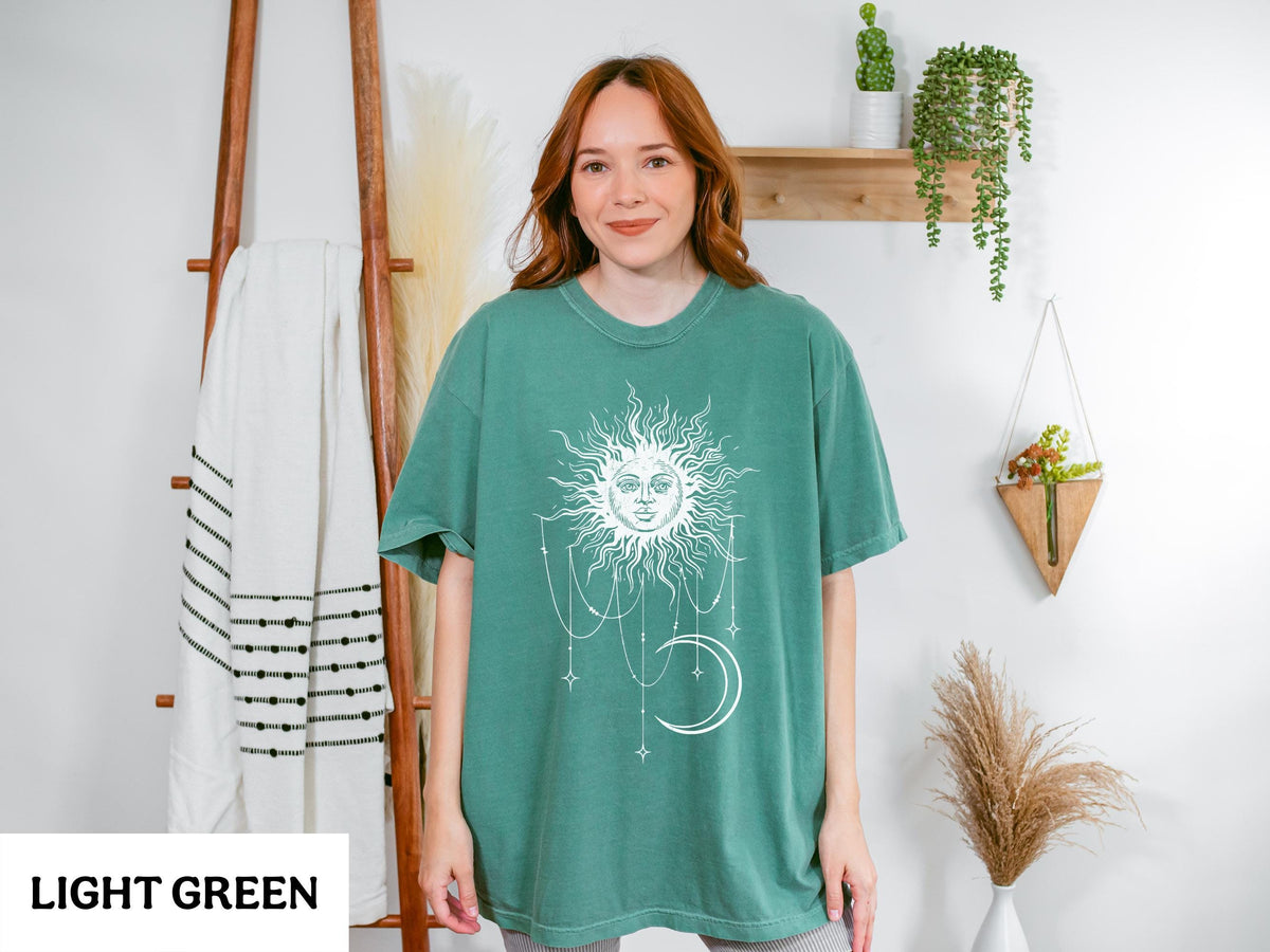 May include: A light green short-sleeve t-shirt featuring a white graphic print of a sun with a human face, surrounded by hanging stars and a crescent moon. The design is centered on the chest of the garment, which has a crew neckline.